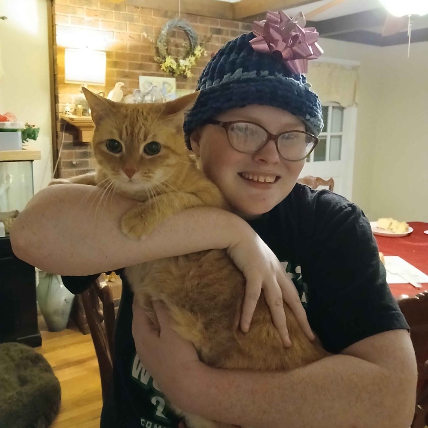 Smiling person wearing a blue hat holds a fluffy orange cat in a cozy indoor setting.