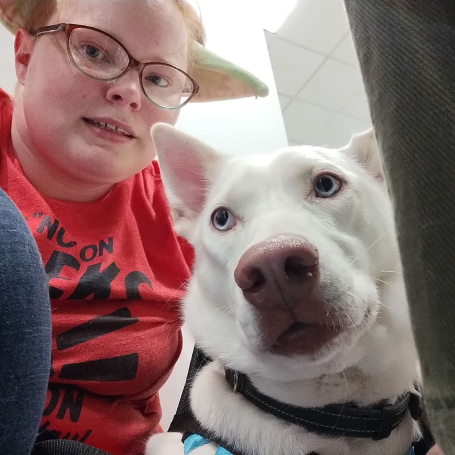 Person with glasses and Yoda ears sits next to a white dog with blue eyes in a casual setting.