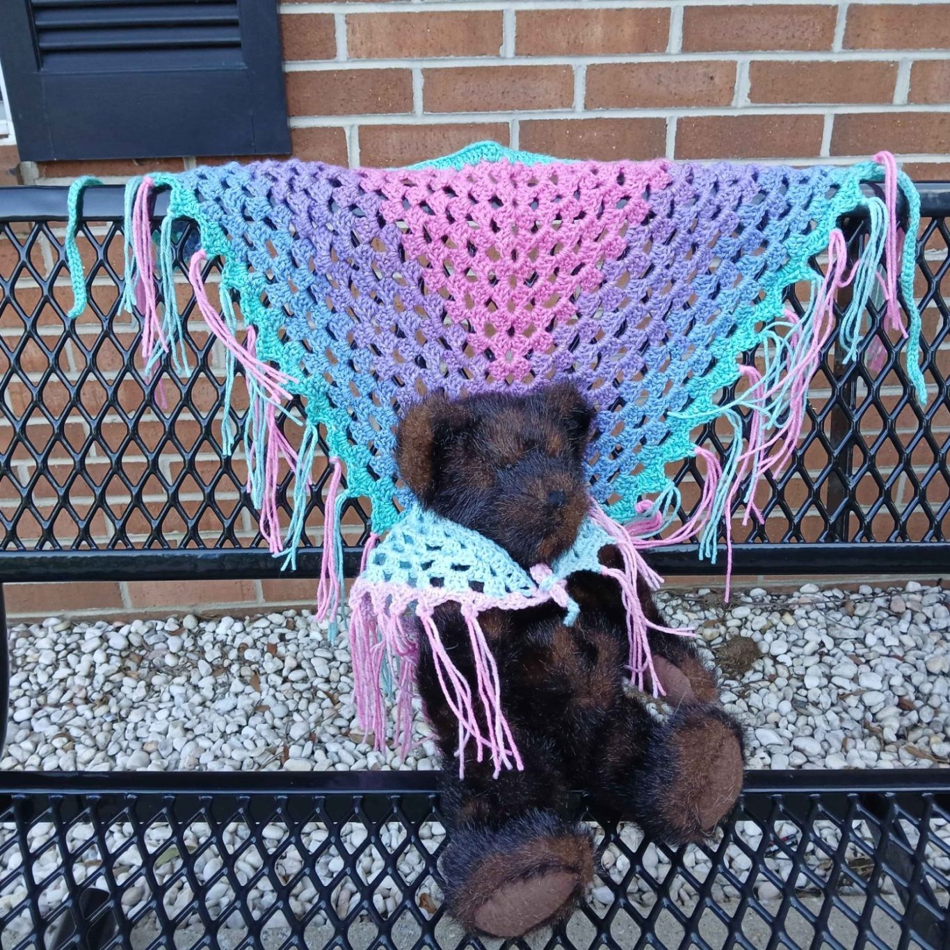 A bear seated on a bench, draped in a colorful, crocheted shawl.